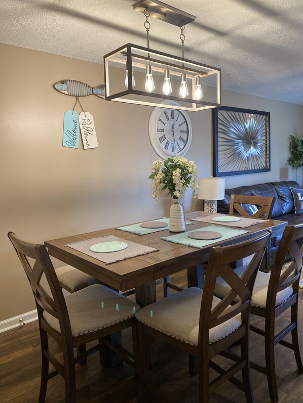 Un espace repas comprenant une table en bois, des chaises rembourrées, un lustre, une horloge murale et des œuvres d’art décoratives accrochées au mur.