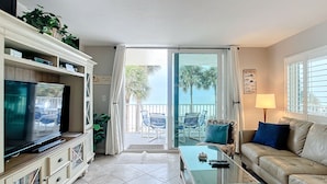 Un salón con televisión, sofá y una mesa de vidrio. Tiene un balcón con sillas y vistas a palmeras.