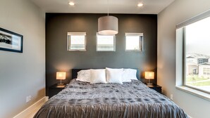 A bedroom with a large bed, two bedside lamps, and three windows. - Downtown Views and Brews (Fort Collins)