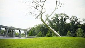 Eine Skulptur eines Baumes in einer grünen Wiese, im Hintergrund ein Gebäude.