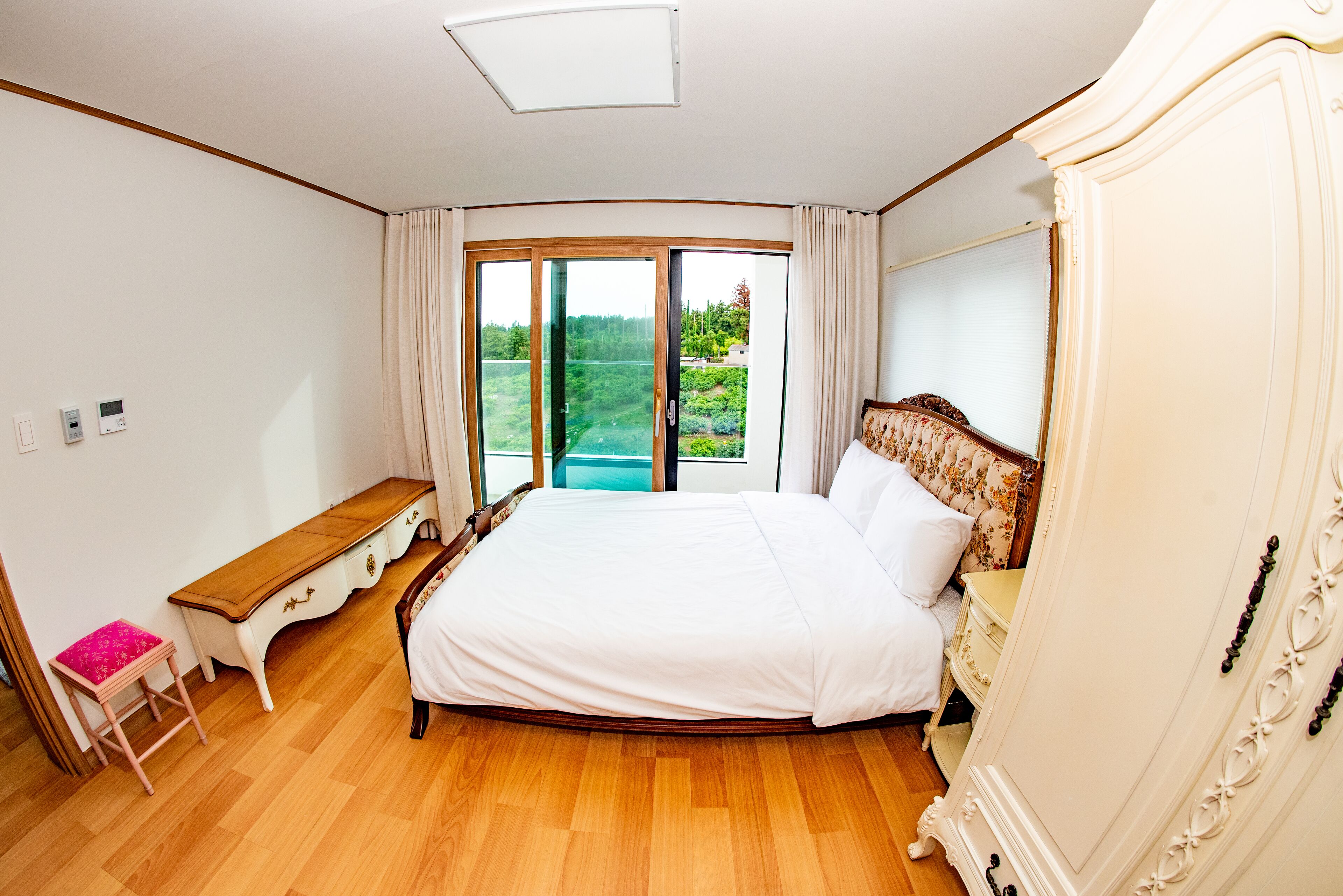 A bedroom with a large bed, a wooden desk, a chair, and a view of a garden through a sliding door.