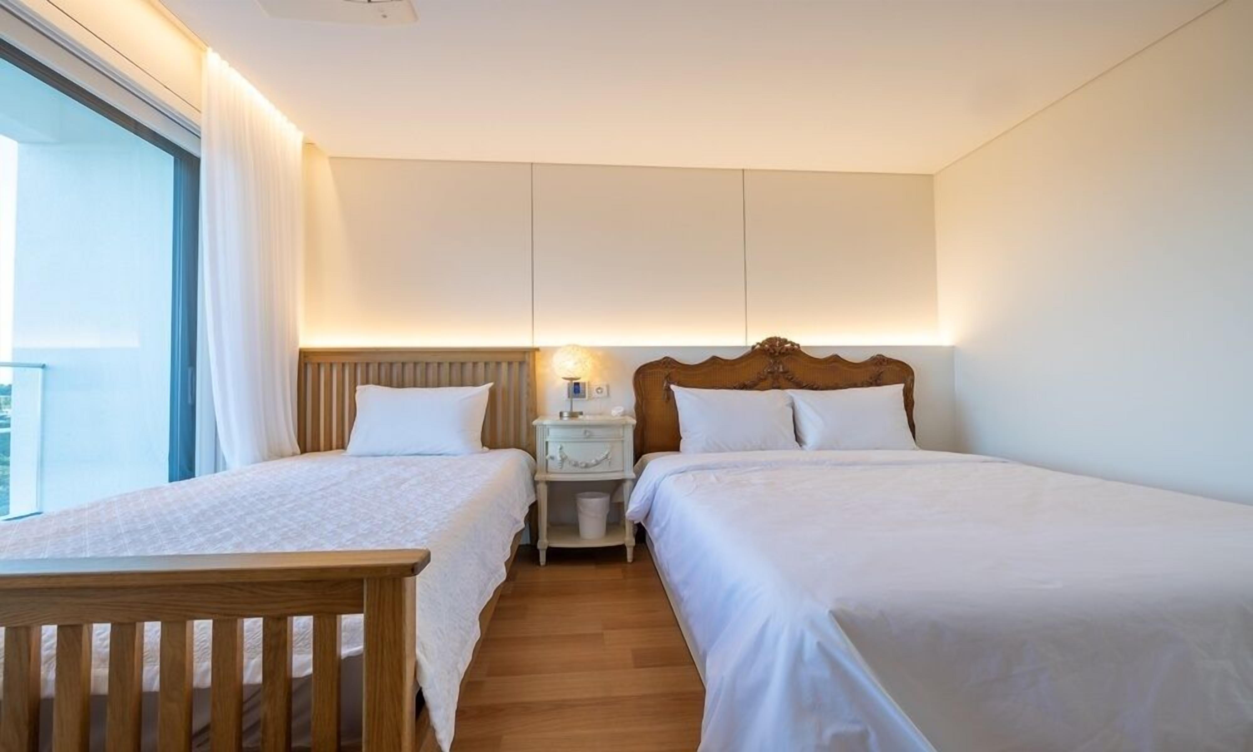 Quarto de hotel com duas camas de solteiro, uma mesa de cabeceira de madeira, um abajur e vista para o exterior.