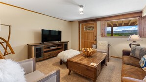 Un soggiorno con televisione, mobili in legno, un pouf bianco e soffice, una lampada e vista sulle montagne attraverso una finestra.