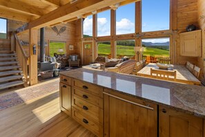 A wooden cabin interior with a kitchen, dining area, and large windows offering a view of the countryside. - Bear Vista (Crested Butte)