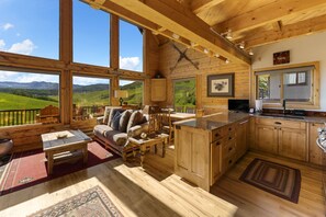 A wooden cabin with a large window, a sofa, a dining table, and a kitchen area. - Bear Vista (Crested Butte)