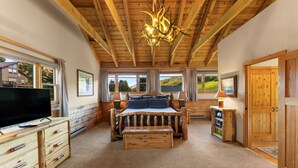 Una camera da letto con soffitto in legno, un letto con copripiumino a righe, una televisione su una cassettiera in legno e una finestra con vista sul verde.