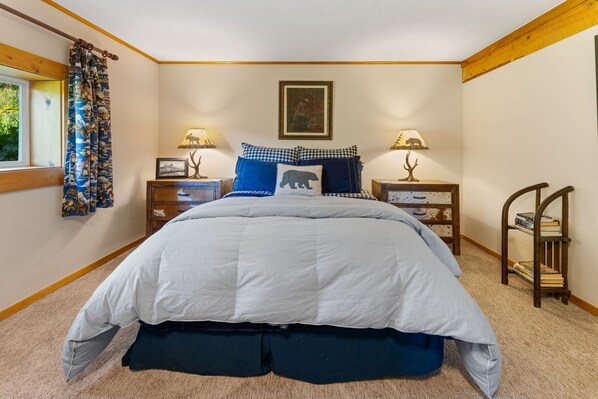 A bedroom with a large bed, two bedside tables with lamps, a window with curtains, a picture on the wall, and a small wooden chair with books.