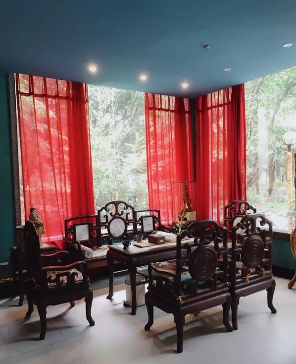 A traditional room with a wooden sofa set, a small table, and a view of a forest through large windows with red curtains.