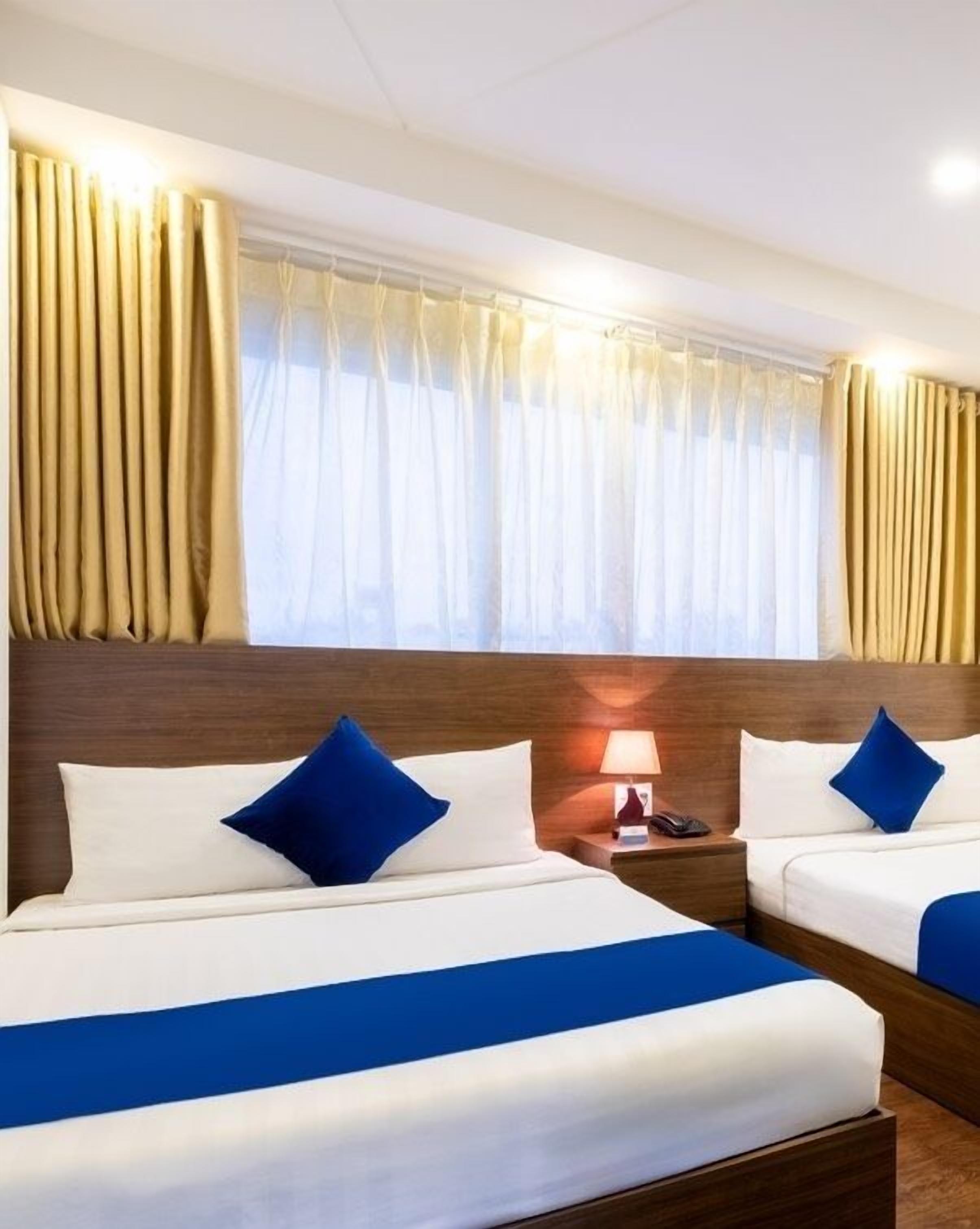 Ein Hotelzimmer mit zwei Betten, jeweils mit weißer und blauer Bettwäsche, einem Holz-Kopfteil, einem Nachttisch mit Lampe und Vorhängen.