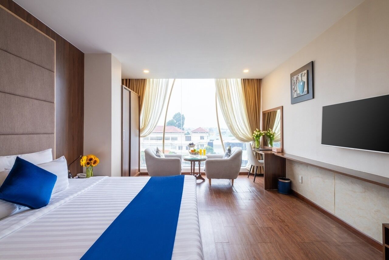 A hotel room with a large bed, a TV, a desk with a chair, and a view of buildings through the window.