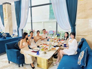 A group of people seated around a table with various dishes, in a room with blue sofas and curtains. - Sandals Hotel (B'Lao)