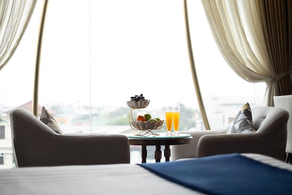 A hotel room with a bed, two armchairs, a small table with a fruit bowl and a glass of orange juice, and a view through large windows.