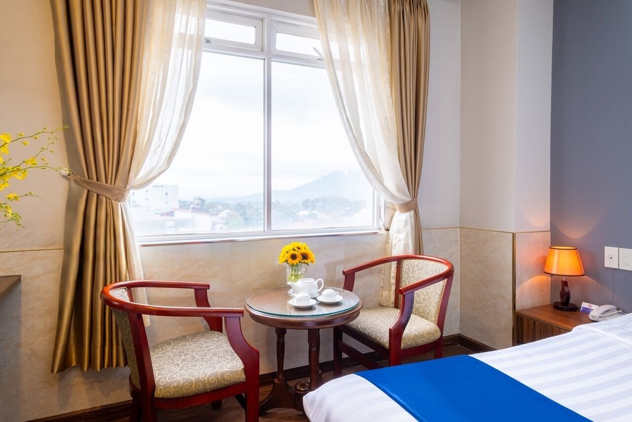 A hotel room with a bed, two chairs, a small table with a vase of sunflowers, a lamp, and a window with curtains.