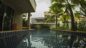 Une piscine dont les bords sont recouverts de carreaux, entourée de verdure et bordée d’un bâtiment doté de grandes fenêtres.