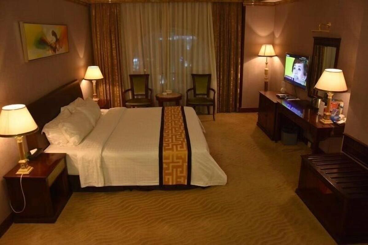A hotel room with a bed, two chairs, a TV, and a desk with a lamp.