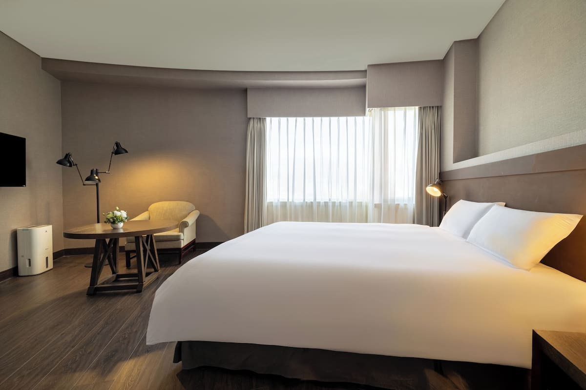 A hotel room with a large bed, a desk with a chair, a lamp, and a window with curtains.