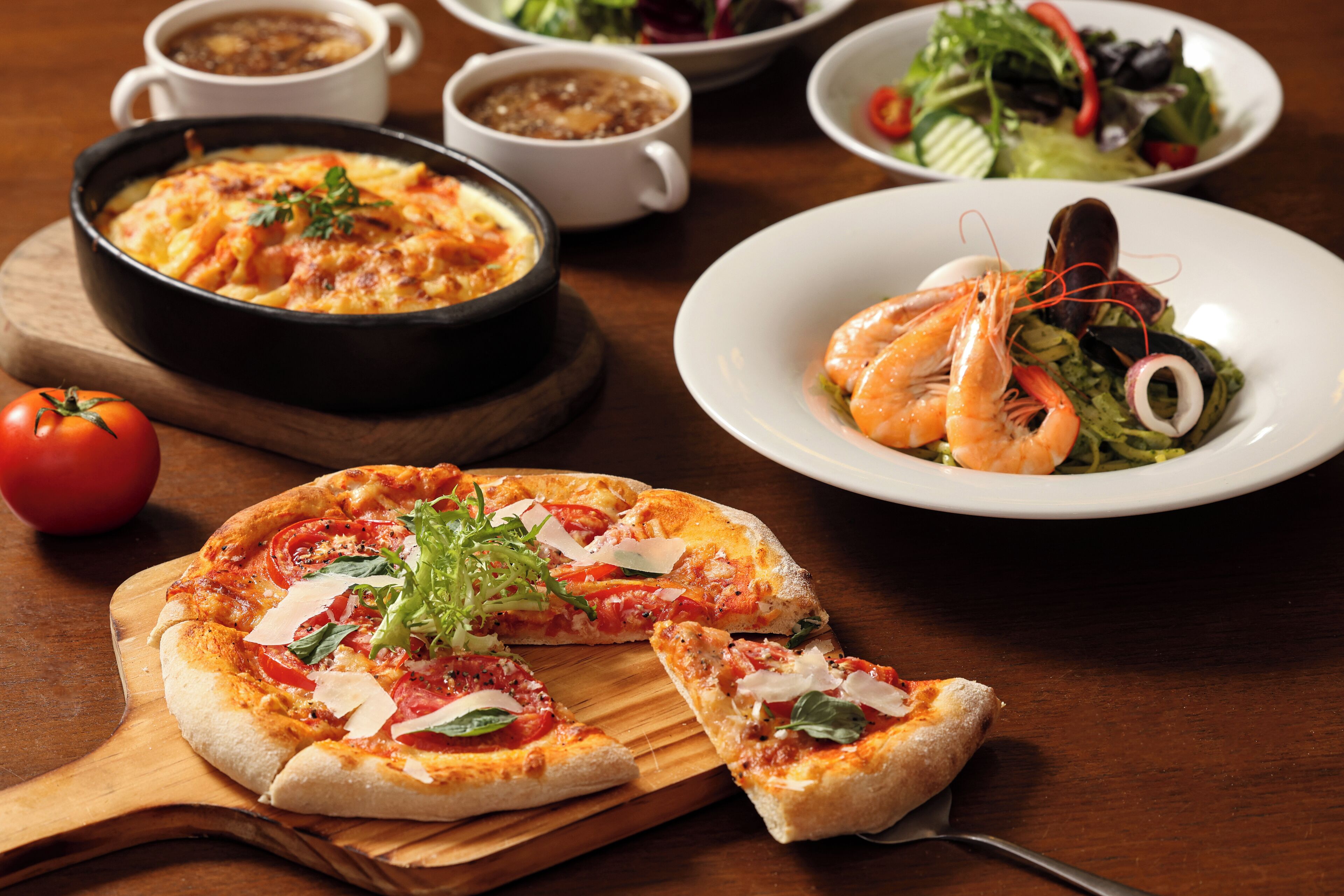 A pizza with toppings, a salad with shrimp, and a bowl of soup on a wooden table.