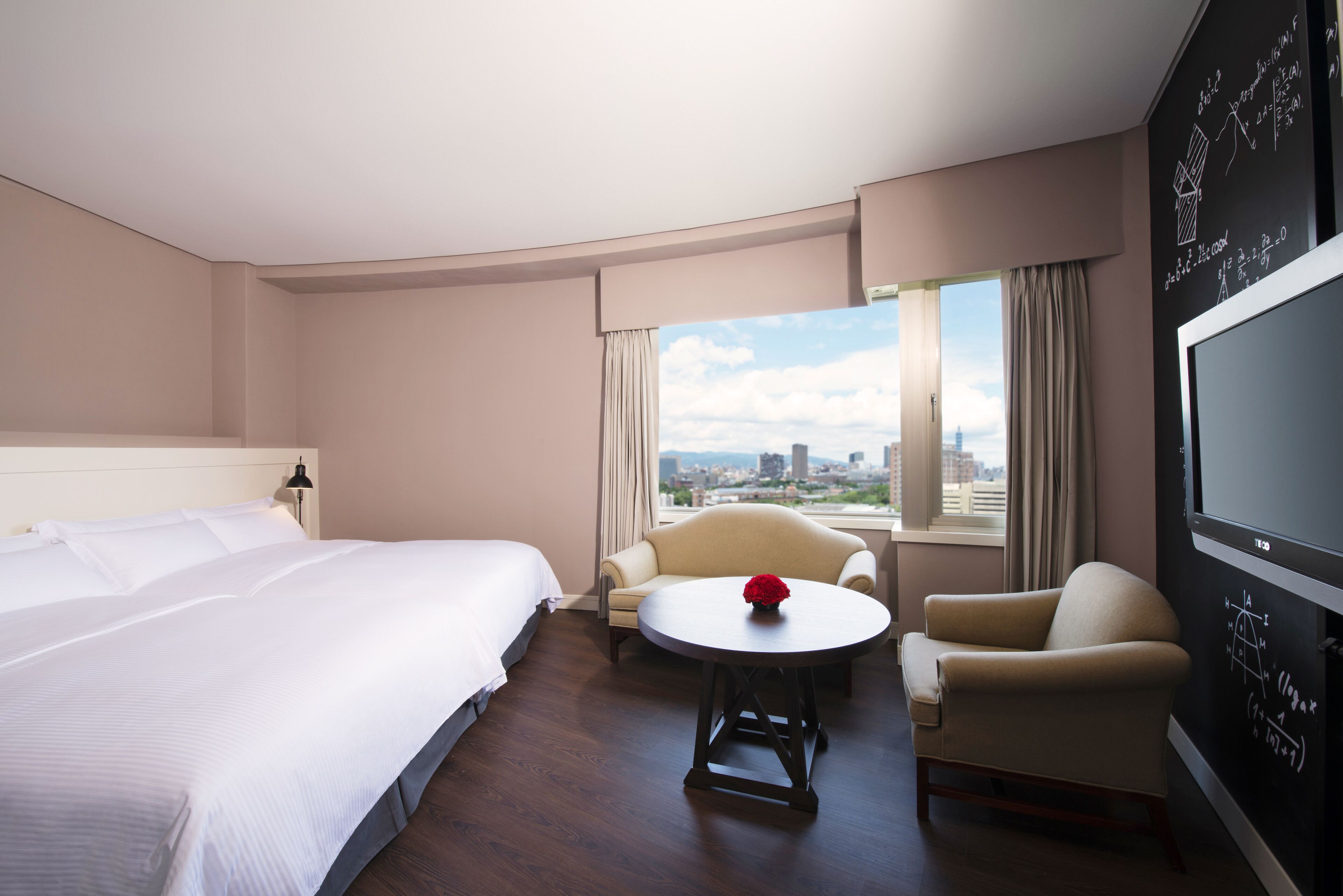 Habitación de hotel con una cama grande, una mesa redonda con una flor roja, un sofá y un televisor en la pared.