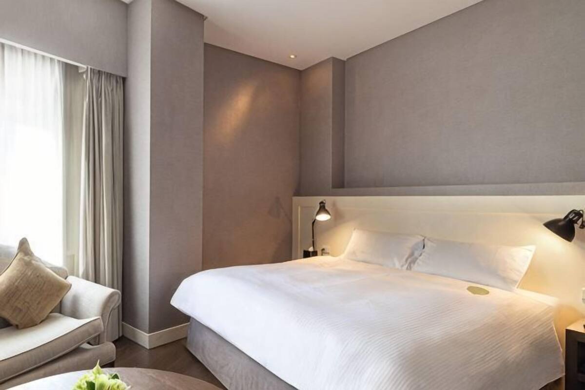 A hotel room with a large bed, a bedside table with a lamp, a sofa, and a window with curtains.