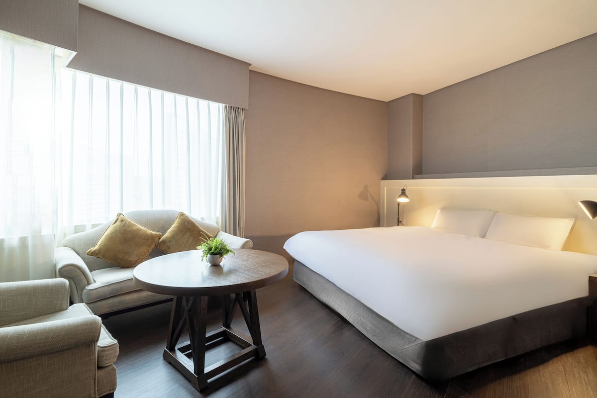 A hotel room with a large bed, a sofa, a round table with a plant, and a window with curtains.