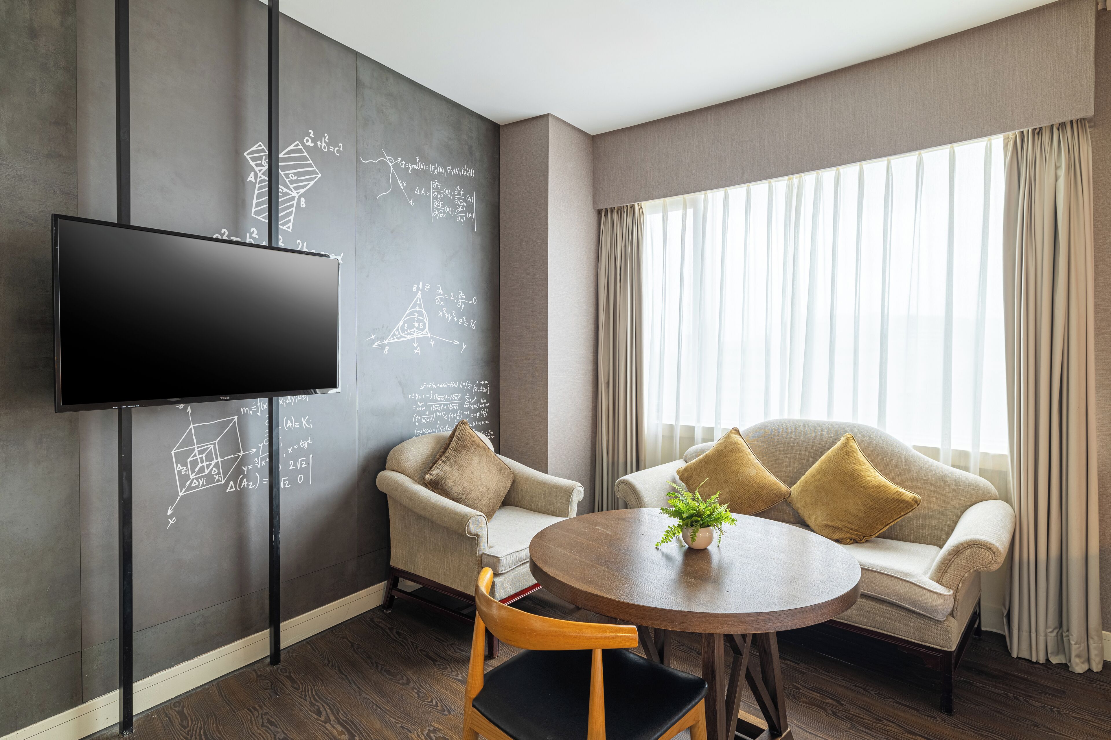 Habitación de hotel moderna con un televisor de pantalla plana grande, un sofá, una silla y una mesa redonda con una planta.