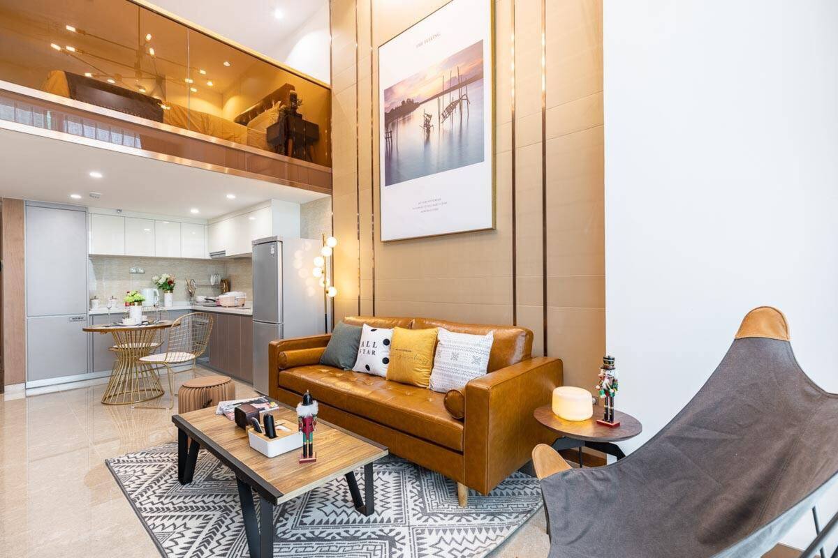 Un salon moderne avec un canapé en cuir marron, une table basse en bois et un fauteuil gris. On aperçoit une cuisine à l’arrière-plan, avec des meubles blancs, ainsi qu’un coin repas comprenant une table ronde et des chaises.