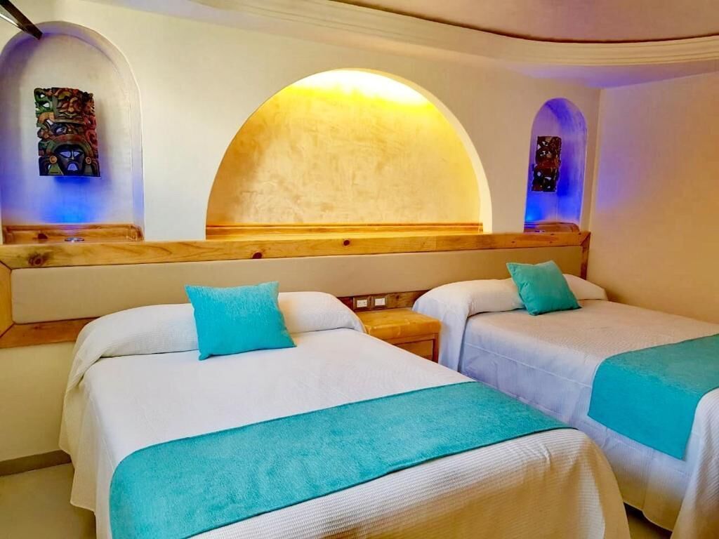 A hotel room with two beds, each with a blue pillow and a matching blue throw blanket.