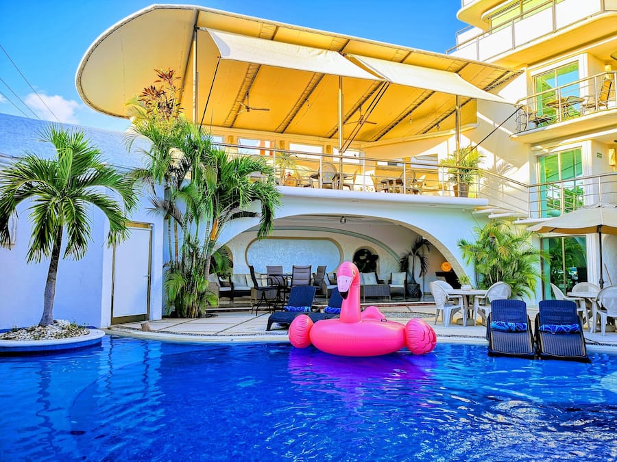Un área de piscina con un flamenco inflable rosa, sillas de descanso y una zona de estar cubierta con mesas y sillas.