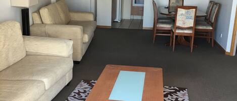 A living room with a beige sofa, a wooden coffee table, and a patterned rug. There is a dining area with a table and chairs, and a kitchen with cabinets and appliances in the background.