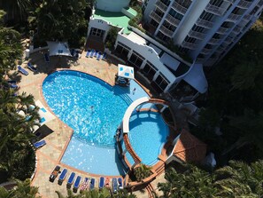 A large, heart-shaped swimming pool surrounded by lounge chairs and palm trees, with a building featuring balconies in the background. - Bell Of Broadbeach (Broadbeach)