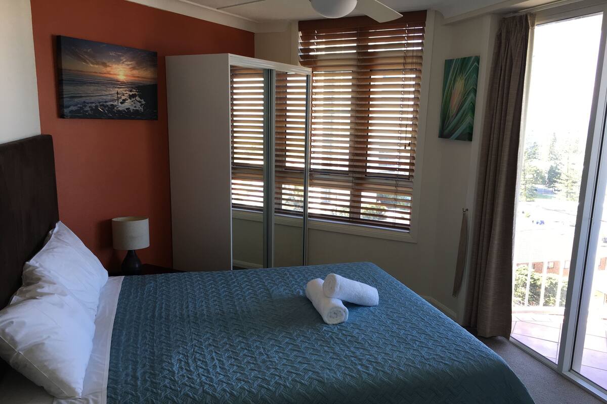A hotel room with a bed, a nightstand, a lamp, a painting, a wardrobe, and a window with blinds.