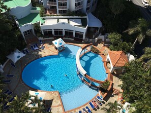 Une grande piscine dotée de nombreux transats et d’un petit toboggan, entourée de palmiers et bordée d’un immeuble à plusieurs étages avec des balcons.