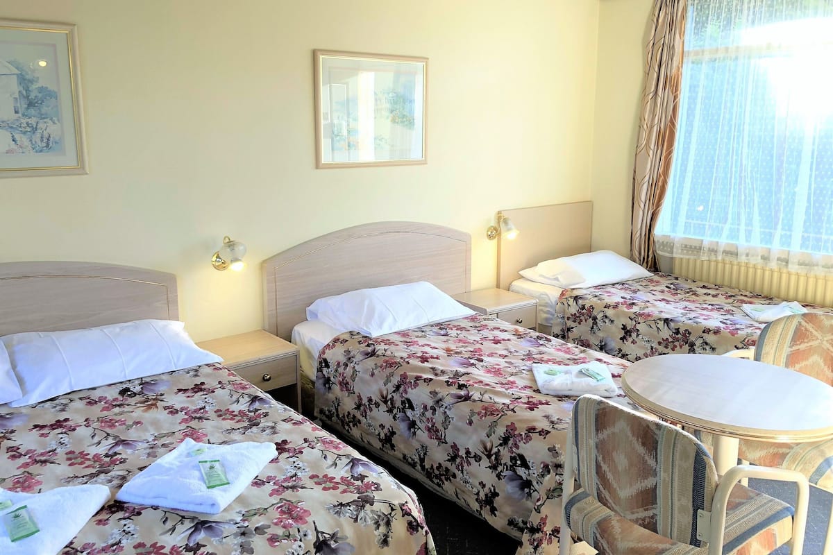 A hotel room with two beds, a small table, and a chair. There are two bedside tables with lamps, a window with curtains, and a framed picture on the wall.