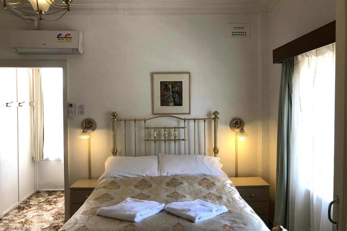 A hotel room with a bed, bedside lamps, a ceiling fan, an air conditioning unit, a framed picture on the wall, and a window with curtains.