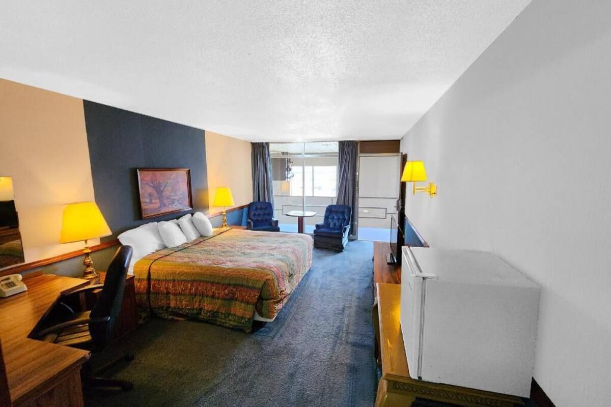 A hotel room with a bed, two chairs, a desk with a lamp, a television, and a refrigerator.