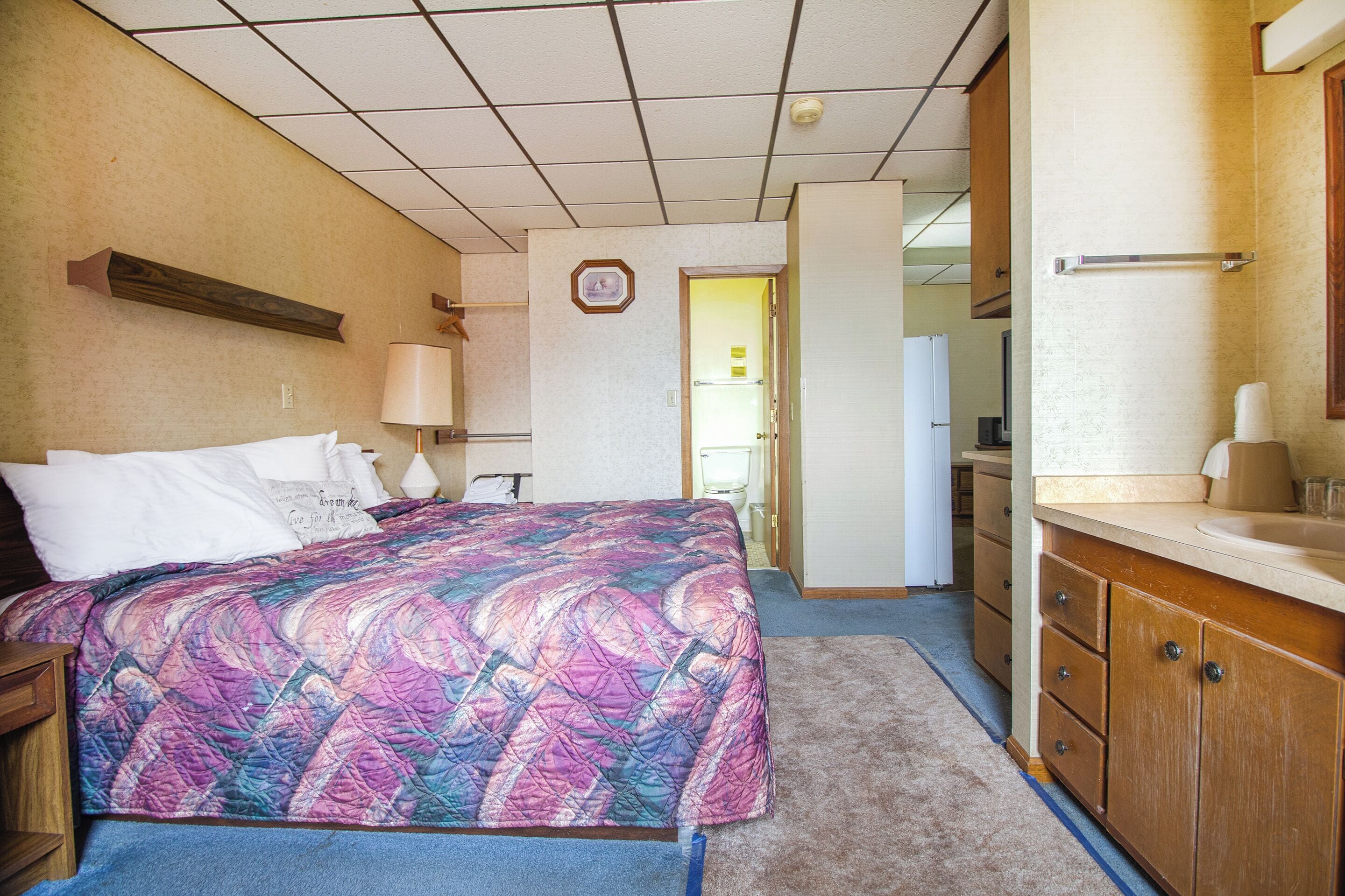 Habitación de hotel con cama, escritorio, lámpara, baño con lavabo y espejo, y una puerta que da a otra habitación.