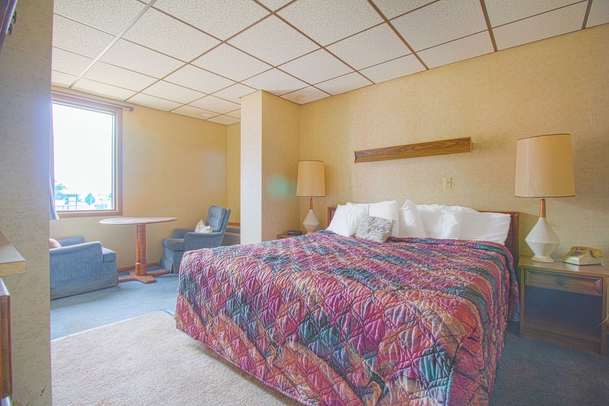 A hotel room with a large bed, two bedside lamps, a small round table, a chair, and a window.