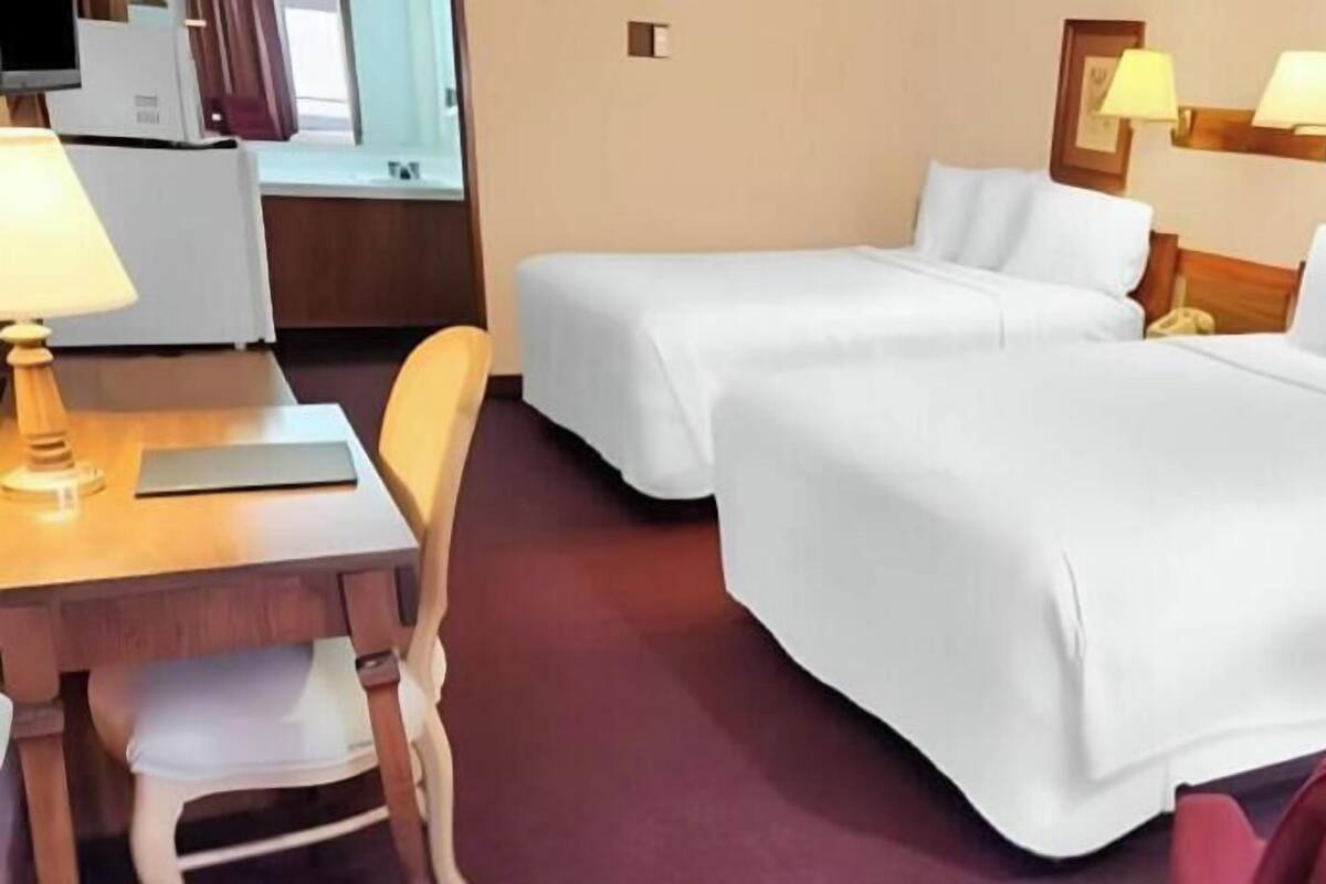 A hotel room with two beds, a desk with a lamp, and a chair.