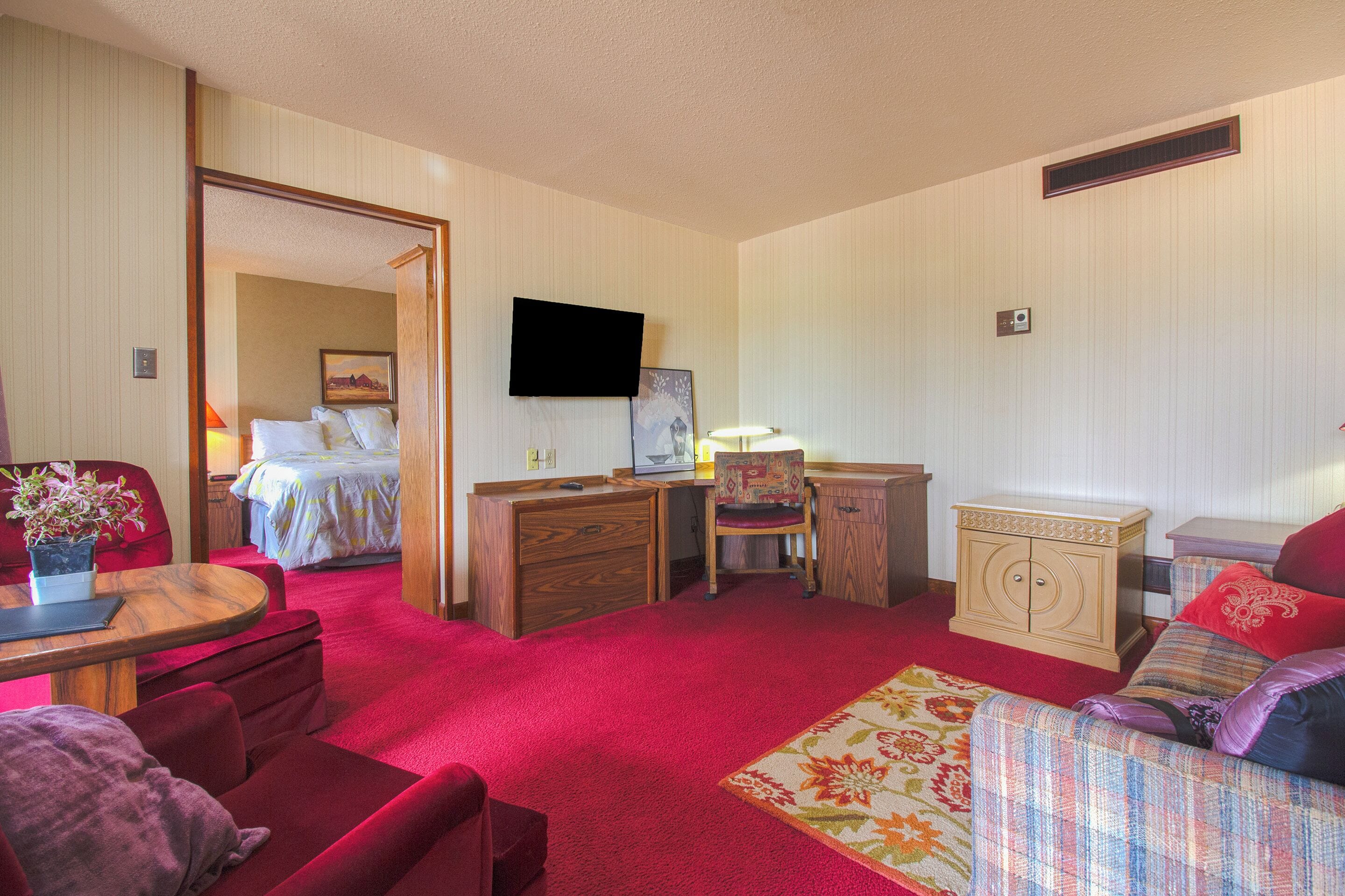 Ein Hotelzimmer mit rotem Teppich, einem Bett mit weißer Bettdecke, einem Holztisch, einem Stuhl, einem an der Wand montierten Fernseher und einem kleinen Schrank.
