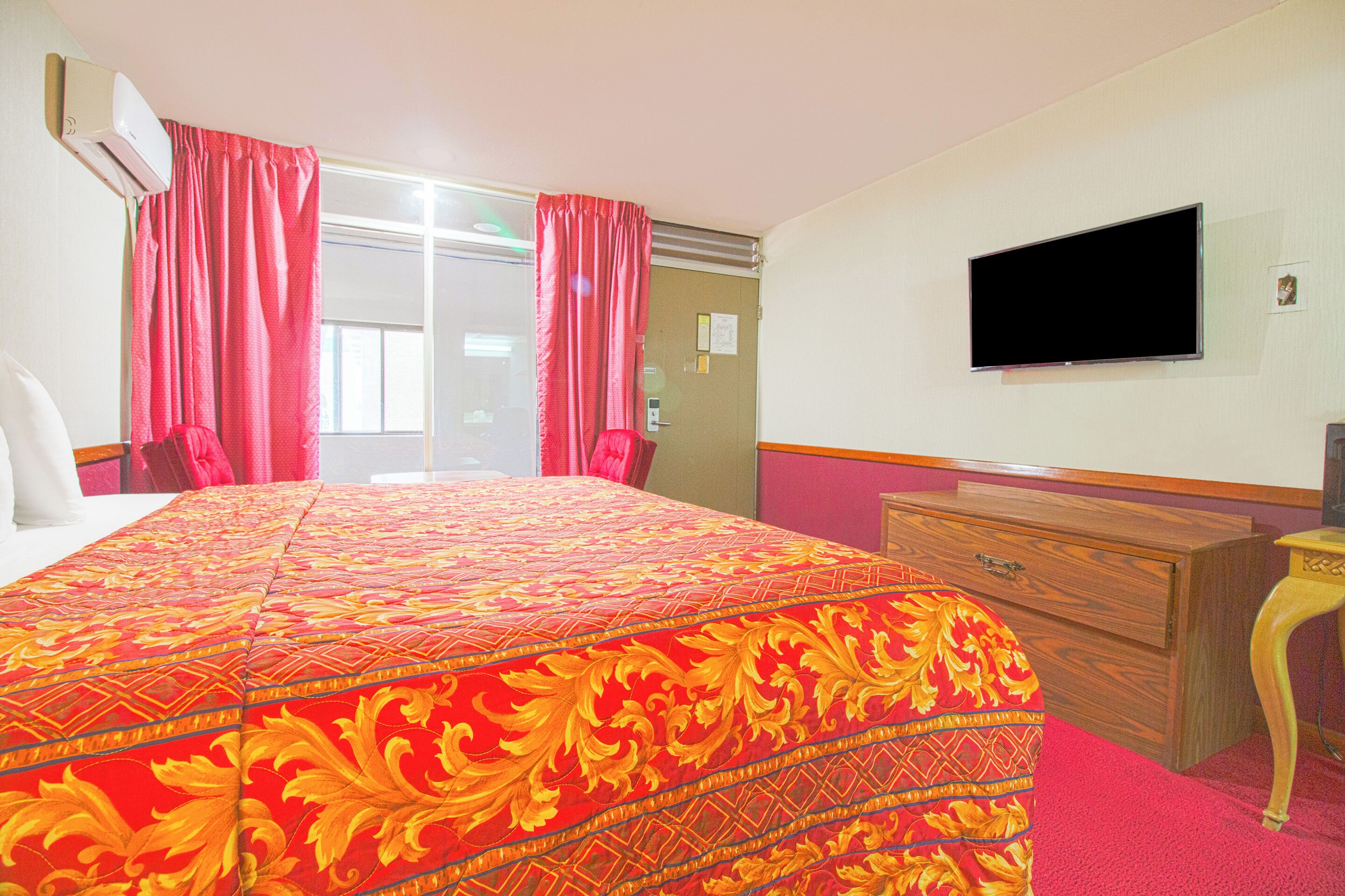 A hotel room with a large bed, a TV on the wall, a wooden dresser, and a window with curtains.