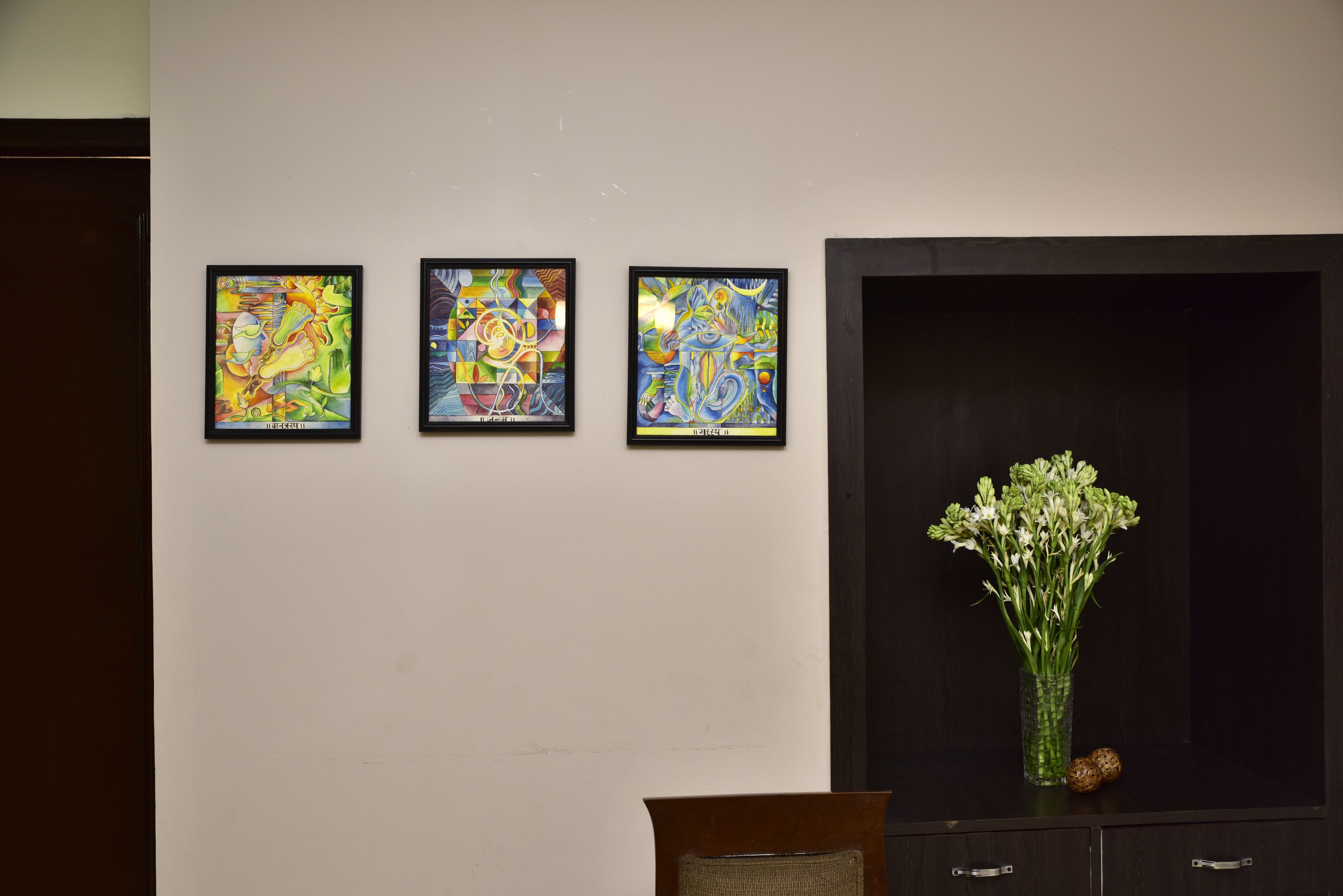 A wall with three framed abstract paintings and a vase with flowers on a cabinet.