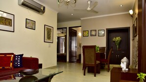 A spacious living area with a sofa, dining table, and chairs. An air conditioning unit is mounted on the wall, and there are framed pictures on the walls. - Woodpecker - 2 Bedroom Service Apartment (New Delhi)