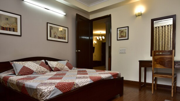 A bedroom with a wooden bed, a desk, and a chair. There are framed pictures on the wall and a mirror above the desk. - Woodpecker - 2 Bedroom Service Apartment (New Delhi)
