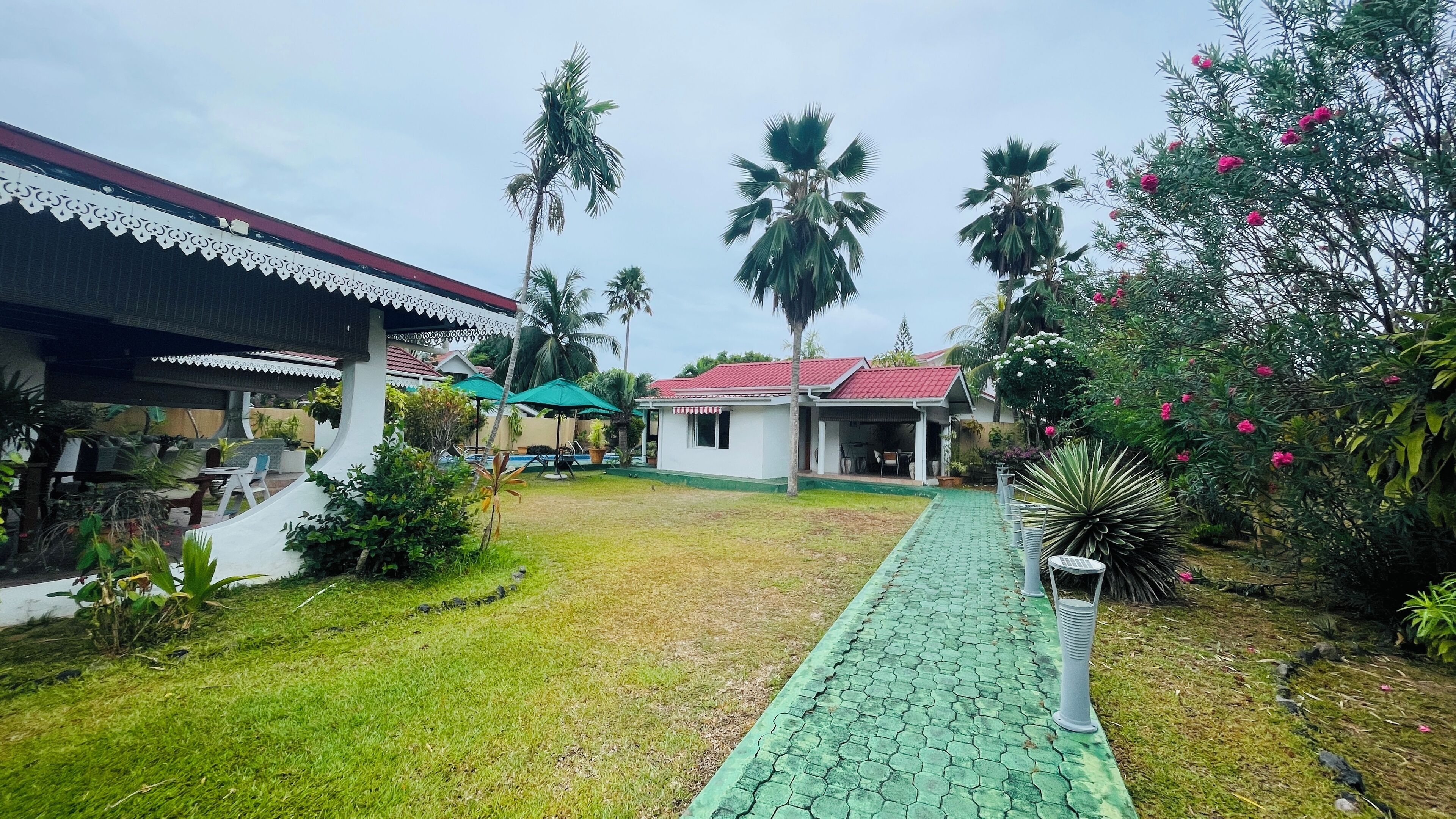 A residential area with a paved walkway, palm trees, and a small house with a red roof.
