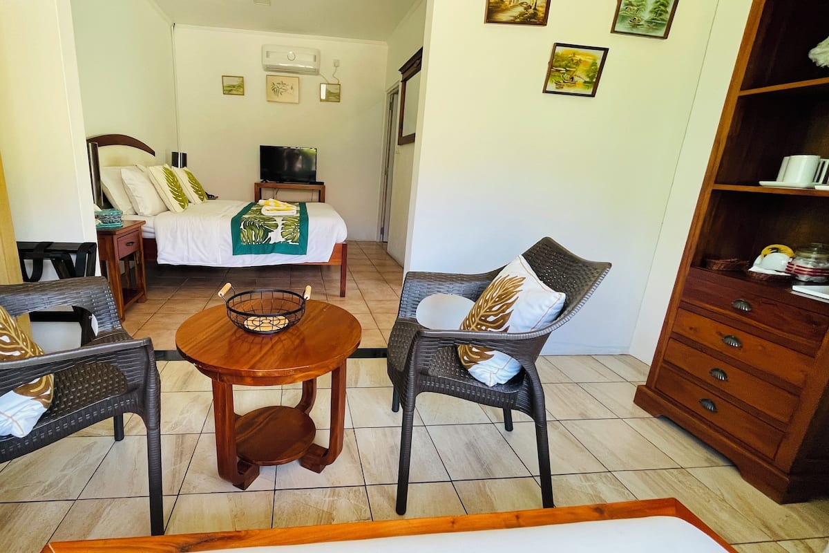 A room with two wicker chairs, a wooden table, a bed with a white bedspread, a small TV on a wooden stand, and a wooden cabinet with drawers.