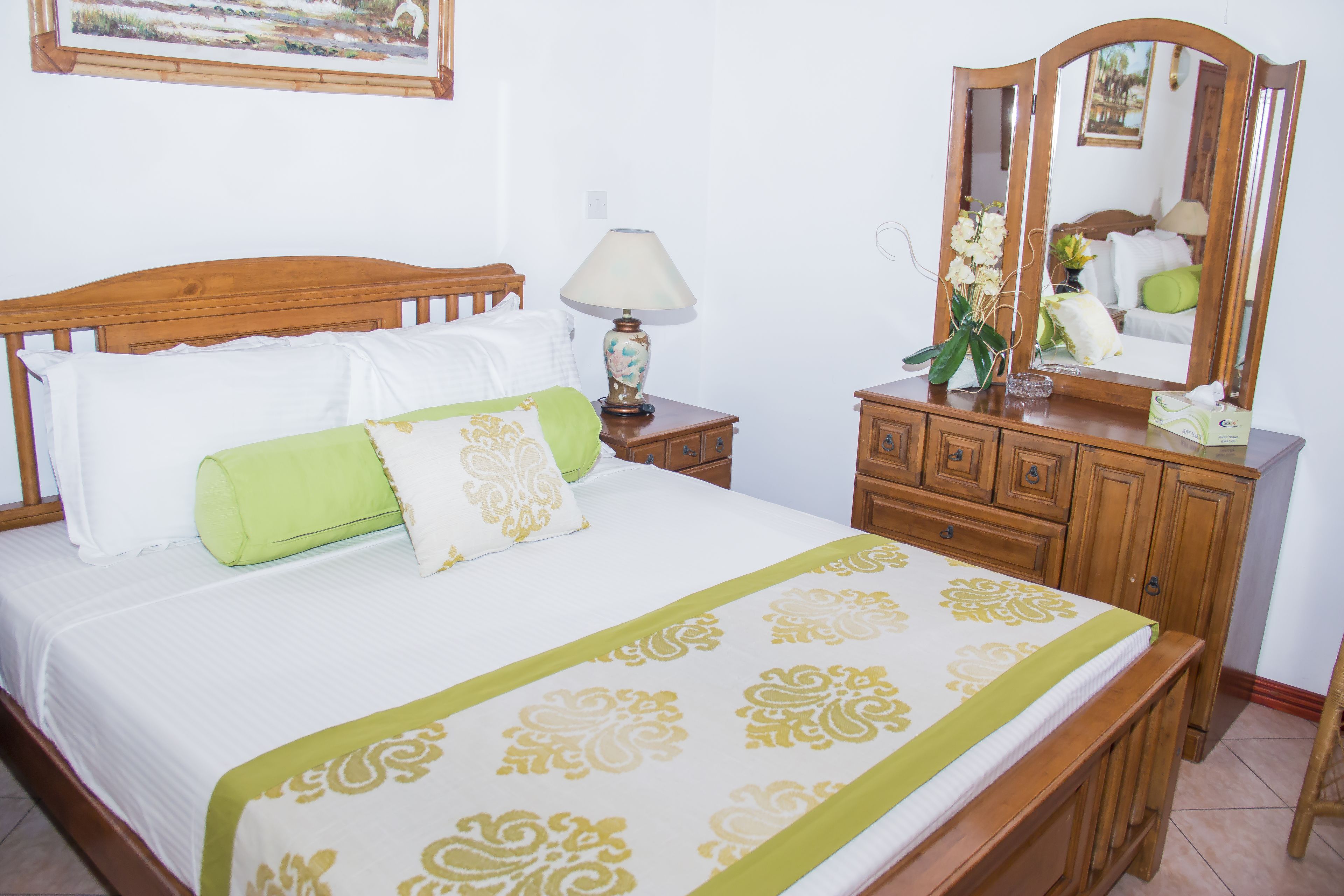 Una cama bien hecha con una colcha verde y blanca, una mesita de noche de madera con una lámpara y un aparador con un espejo.