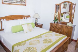 Una cama bien hecha con una colcha verde y blanca, una mesita de noche de madera con una lámpara y un aparador con un espejo.