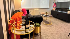 En moderne stue med en sort sofa, et rundt guld bord med vinglas og en flaske, samt et fjernsyn på et sort skab.