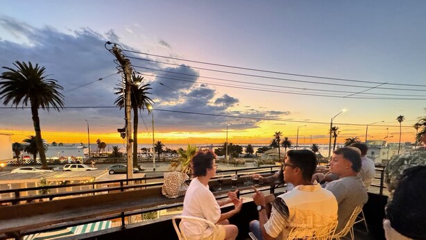 En terrasse med metalstole og borde, med udsigt over en solnedgang med palmer og en strand i det fjerne.