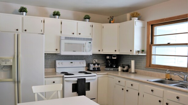 Una cocina con armarios blancos, microondas, estufa, fregadero y una ventana con persianas.